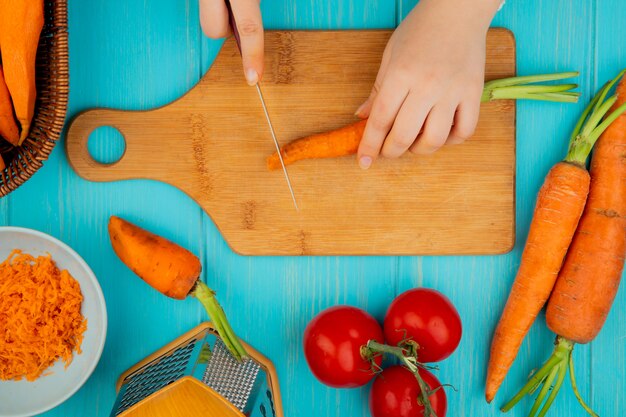 Vista superior de manos de mujer cortando zanahoria en tabla de cortar con tomate tomate sobre fondo azul.