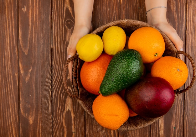 Vista superior de las manos de mujer sosteniendo una cesta llena de cítricos como aguacate mango limón naranja sobre mesa de madera con espacio de copia