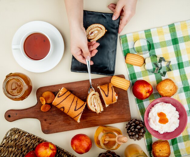 Vista superior de las manos de mujer sosteniendo una rodaja con un tenedor en la tabla de cortar con ciruelas secas, duraznos, mermeladas, requesón, galletas y piñas y té alrededor en la mesa blanca