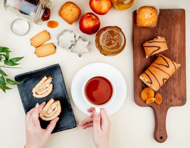 Vista superior de manos de mujer sosteniendo una taza de té y una rebanada de rollo con mermelada, galletas, pasas y ciruelas secas en mesa blanca