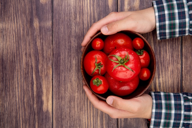 Vista superior de manos de mujer sosteniendo un tazón de tomates en la mesa de madera