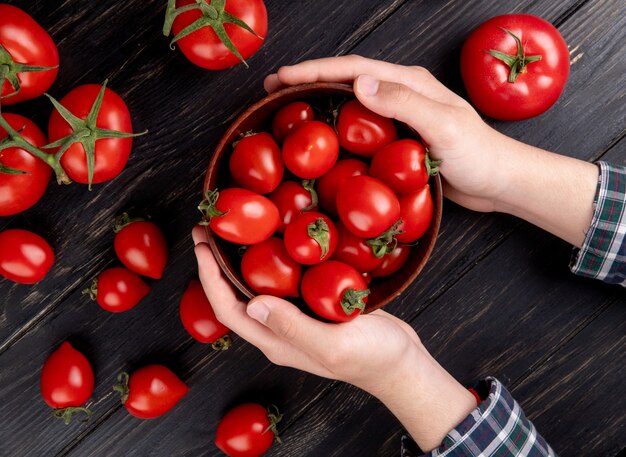 Vista superior de manos de mujer sosteniendo un tazón de tomates con otros en la mesa de madera