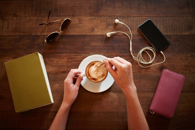 Vista superior de manos recortadas revolviendo capuchino con gafas, libro, billetera y teléfono inteligente acostado sobre la mesa