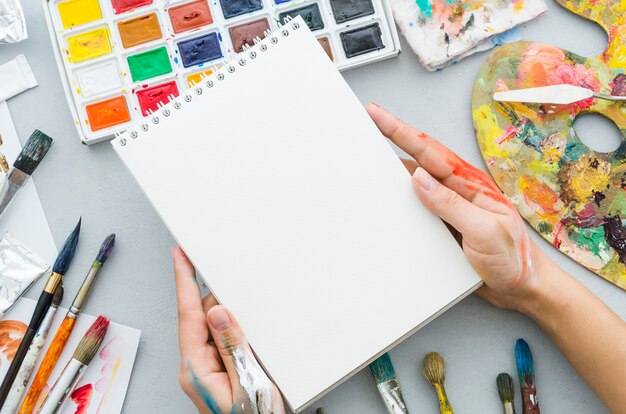 Vista superior manos sosteniendo cuaderno rodeado por elementos de pintura