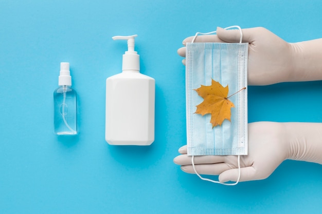 Vista superior de manos sosteniendo mascarilla médica con hoja de otoño y botella de jabón líquido