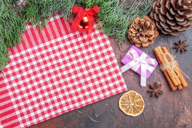 Vista superior mantel a cuadros rojo y blanco ramas de pino piñas regalo de navidad juguete de bola de árbol de navidad de canela sobre fondo rojo oscuro