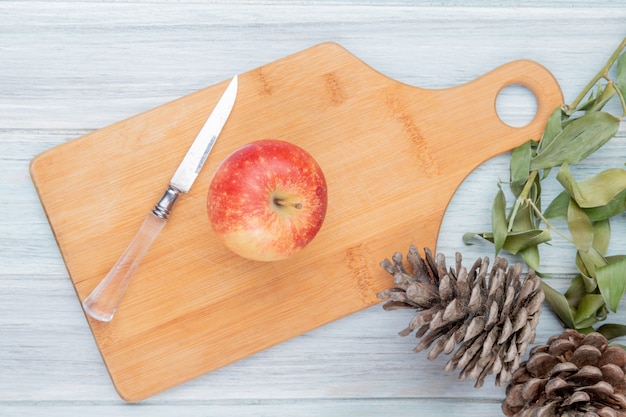 Vista superior de manzana roja y cuchillo en tabla de cortar con piñas y hojas sobre fondo de madera