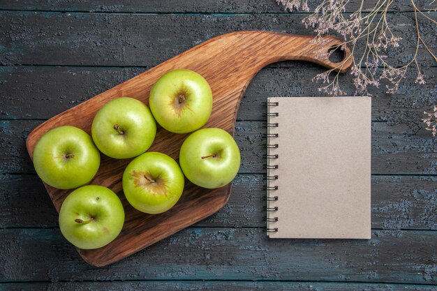 Vista superior de las manzanas a bordo de seis manzanas verdes en el tablero de la cocina junto a las ramas de los árboles y un cuaderno gris sobre una superficie oscura