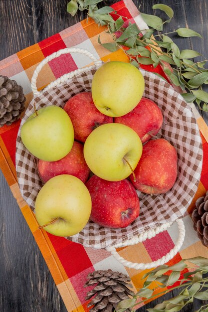 Vista superior de manzanas en canasta con piñas y hojas sobre tela escocesa y madera