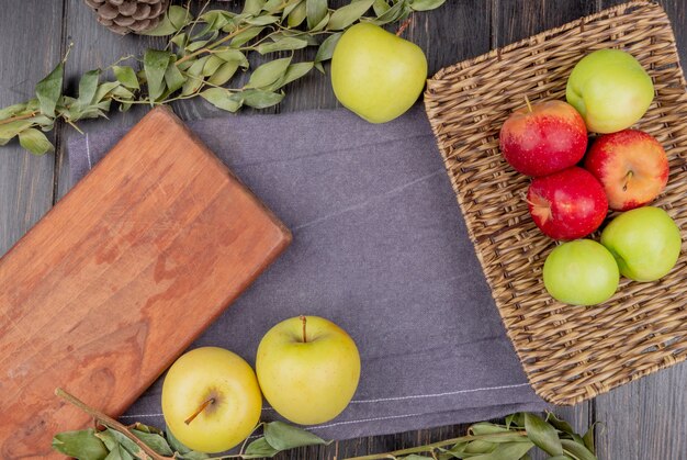 Vista superior de manzanas en canasta y en tela gris con tabla de cortar y hojas en la mesa de madera