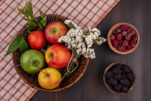 Vista superior de manzanas de colores en una canasta con flores blancas, frambuesas y moras sobre una superficie de madera