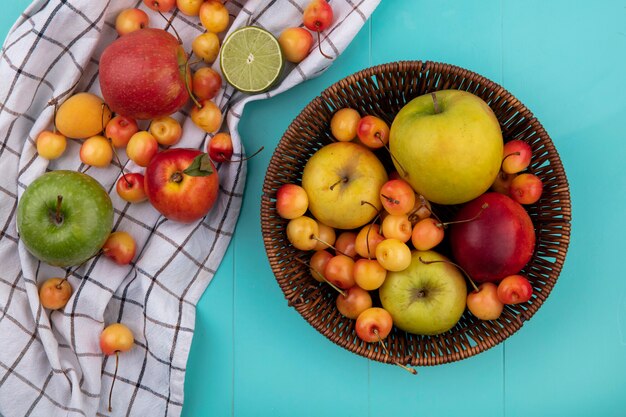 Vista superior de manzanas de colores con cerezas blancas en una canasta sobre una toalla a cuadros sobre una superficie turquesa