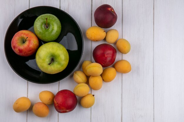 Vista superior de manzanas de colores sobre una placa negra con albaricoques y melocotones sobre una superficie blanca