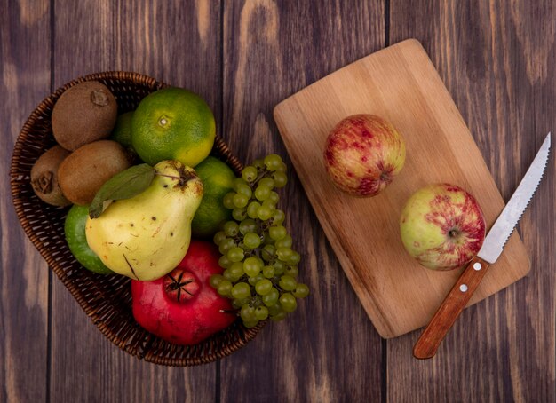 Vista superior de las manzanas con un cuchillo en la tabla de cortar con uvas, mandarinas, pera y granada en la canasta en la pared de madera