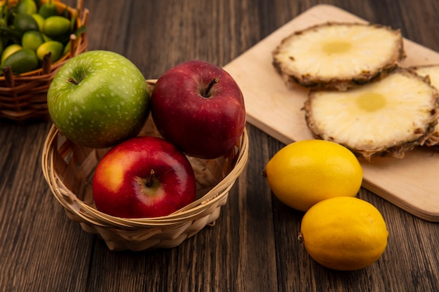 Vista superior de manzanas frescas de colores en un balde con piñas en una tabla de cocina de madera con kinkans en balde con limones aislado en una pared de madera