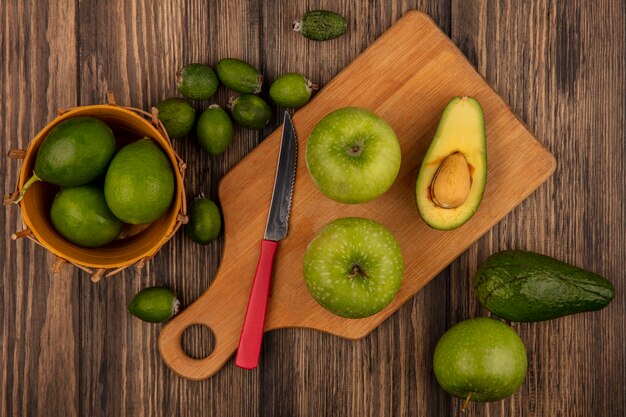 Vista superior de manzanas frescas sobre una tabla de cocina de madera con cuchillo con limas en un balde con feijoas y aguacates aislado sobre un fondo de madera