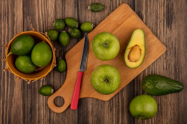 Vista superior de manzanas frescas sobre una tabla de cocina de madera con cuchillo con limas en un balde con feijoas y aguacates aislado sobre un fondo de madera