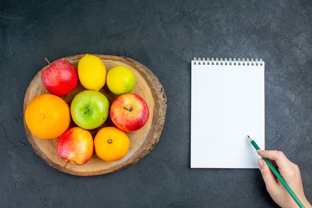 Vista superior manzanas naranjas limón sobre tablero de madera lápiz de cuaderno en mano femenina sobre superficie oscura