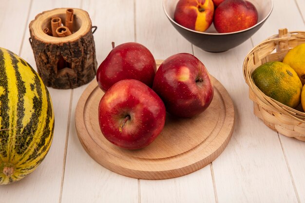 Vista superior de manzanas rojas frescas en una tabla de cocina de madera con melón cantalupo con mandarinas en un cubo sobre un fondo de madera blanca