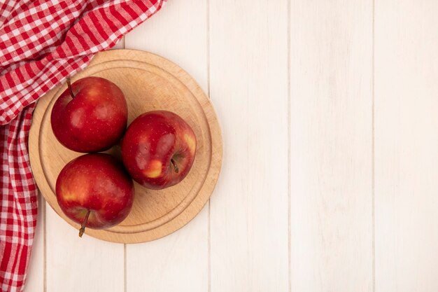 Vista superior de manzanas rojas sobre una tabla de cocina de madera sobre un paño rojo marcado sobre una superficie de madera blanca con espacio de copia