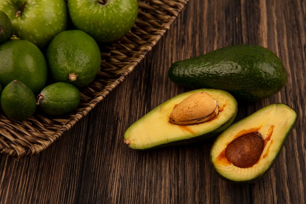 Vista superior de manzanas verdes en una bandeja de mimbre con feijoas y limas con aguacates aislado sobre un fondo de madera