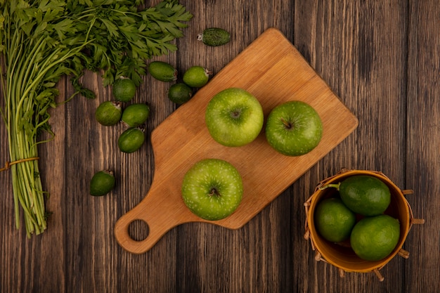 Vista superior de manzanas verdes frescas en una tabla de cocina de madera con limas en un balde con feijoas y perejil aislado en una pared de madera