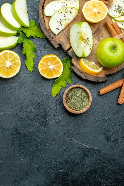 Vista superior de manzanas verdes, palitos de canela y rodajas de limón y manzana en tablero de madera, limones cortados en mesa negra