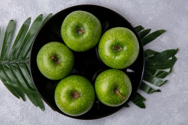 Vista superior de manzanas verdes en un plato negro en ramas con hojas sobre un fondo blanco.