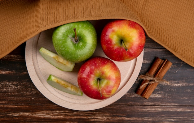 Vista superior de manzanas verdes y rojas en un soporte con canela y una toalla marrón sobre un fondo de madera