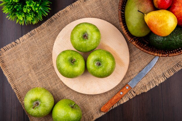 Vista superior de manzanas verdes en una tabla de cocina de madera con un cuchillo sobre tela de saco con un cubo de frutas en la superficie de madera