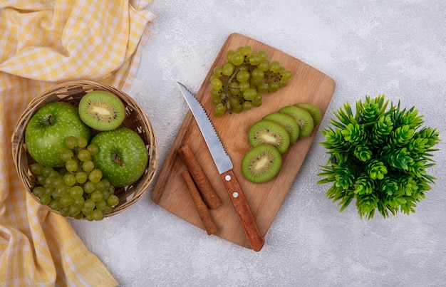 Vista superior de manzanas verdes con uvas verdes y una rodaja de kiwi en una canasta con una toalla a cuadros amarilla y canela con un cuchillo sobre una tabla de cortar sobre un fondo blanco.