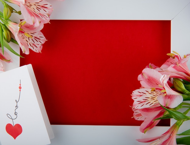 Foto gratuita vista superior de un marco blanco vacío con flores de color rosa alstroemeria y una postal sobre fondo rojo con espacio de copia