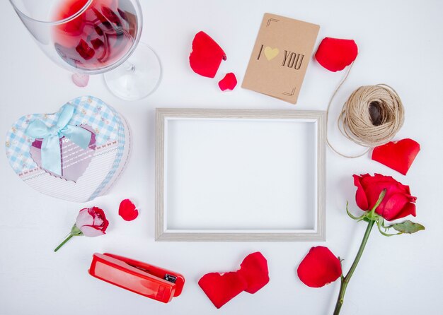 Vista superior de un marco de imagen vacío con una caja de regalo vaso de vino bola de cuerda rosas de color rojo pequeña grapadora postal sobre fondo blanco