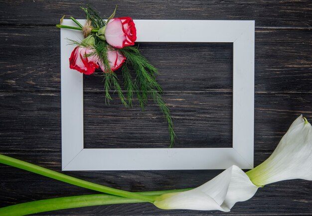 Vista superior de un marco de imagen vacío con rosas rojas con hinojo y alcatraces de color blanco sobre fondo de madera oscura con espacio de copia