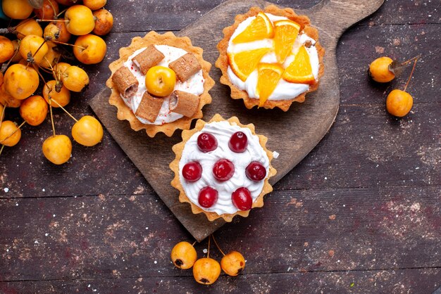 Vista superior más cercana de pequeños pasteles deliciosos con crema de frutas en rodajas y cerezas amarillas frescas en marrón