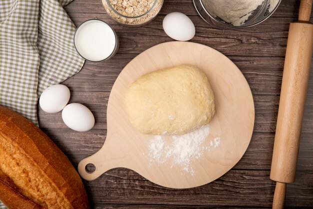 Vista superior de masa con harina en la tabla de cortar y huevos baguette leche rodillo sobre fondo de madera