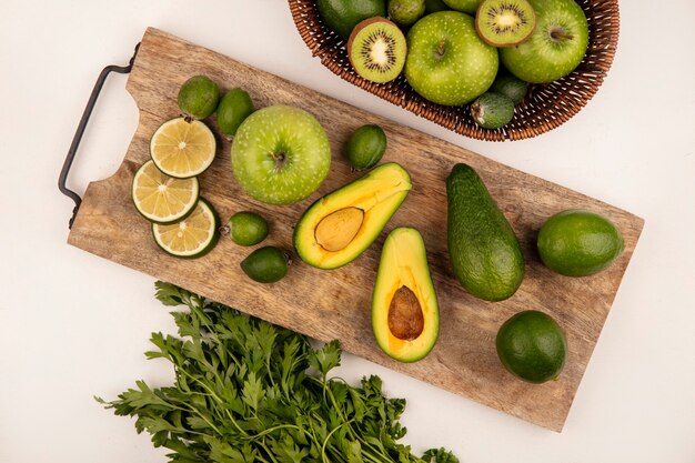 Vista superior de medio aguacate aislado en una tabla de cocina de madera con limas y feijoas con un balde de kiwis y manzanas sobre una superficie blanca