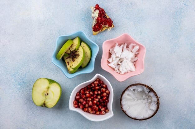 Vista superior de medio coco fresco con pulpas de coco en un tazón rosa rodajas de manzana y semillas de granada en un tazón blanco sobre superficie blanca