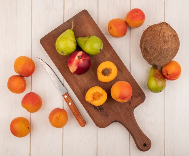 Vista superior de medio corte y frutas enteras como melocotón albaricoques y peras en tabla de cortar y patrón o albaricoques pera y coco y cuchillo sobre fondo de madera