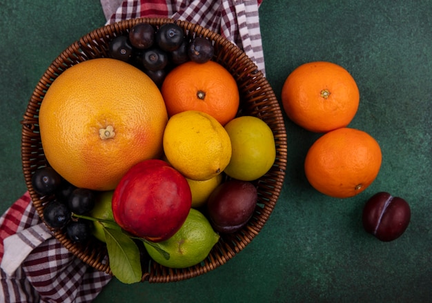 Vista superior melocotón con ciruelas cereza naranjas limones limas y pomelo en una canasta con una toalla a cuadros sobre un fondo verde