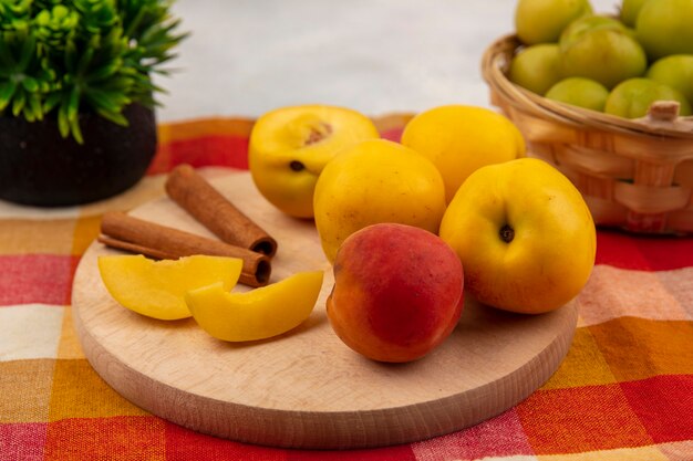 Vista superior de melocotones amarillos en una tabla de cocina de madera con ramas de canela con ciruelas cereza verde en un cubo sobre un fondo de mantel a cuadros