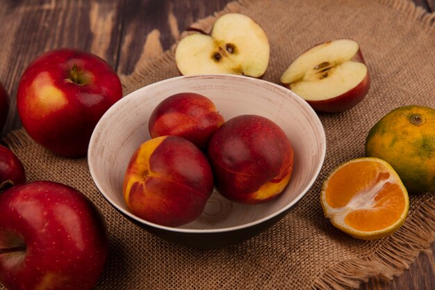 Vista superior de melocotones jugosos en un recipiente sobre una tela de saco con manzanas y mandarinas aislado en una pared de madera