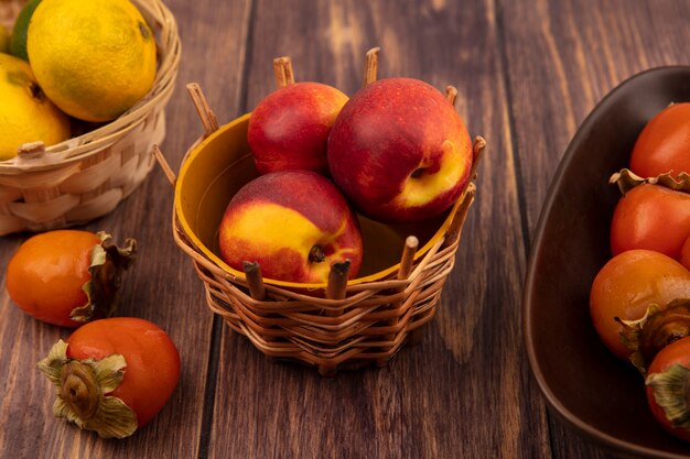 Vista superior de melocotones orgánicos en un balde con mandarinas con caquis aislado en una pared de madera