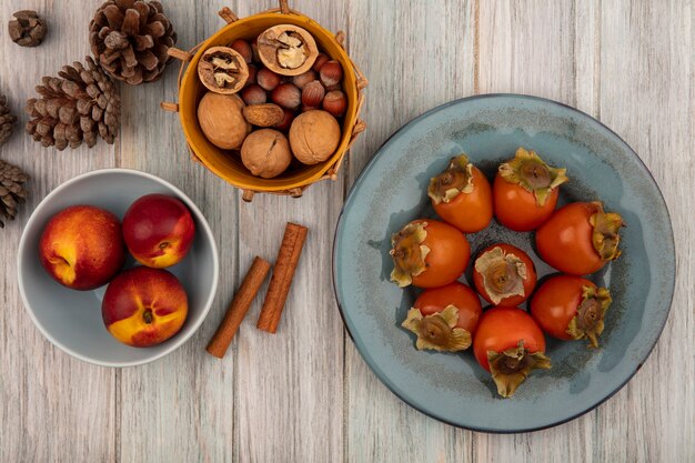 Foto gratuita vista superior de melocotones en un recipiente con caquis en un plato con ramas de canela y nueces en un balde sobre una superficie de madera gris