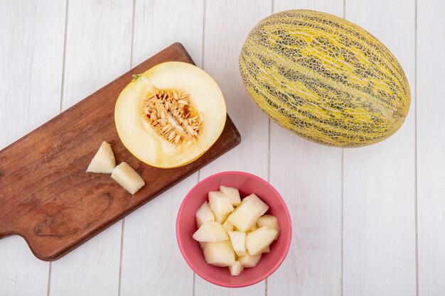 Vista superior de melón sobre una tabla de cocina de madera con rodajas en un recipiente sobre una superficie de madera blanca