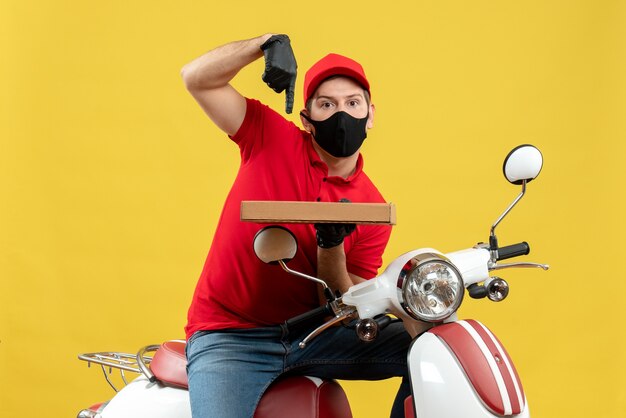 Vista superior del mensajero ambicioso emocional hombre vestido con blusa roja y guantes de sombrero en máscara médica sentado en scooter apuntando orden