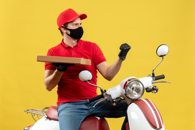 Vista superior del mensajero ambicioso orgulloso emocional hombre vestido con blusa roja y guantes de sombrero en máscara médica sentado en scooter mostrando orden