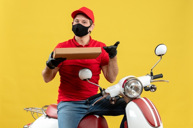 Vista superior del mensajero confiado con blusa roja y guantes de sombrero en máscara médica sentado en scooter mostrando orden