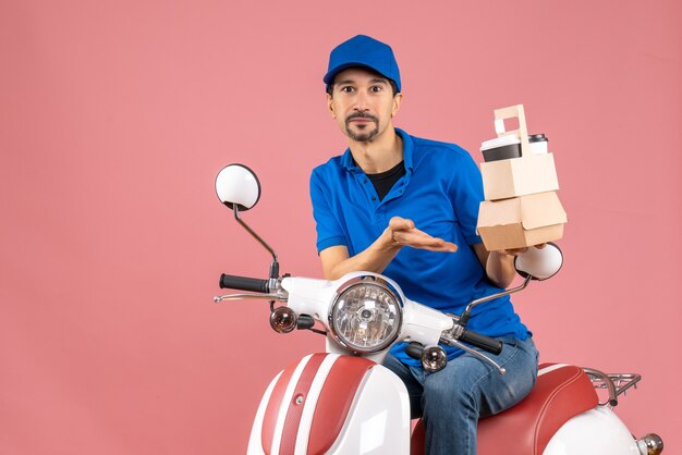 Vista superior del mensajero confiado con sombrero sentado en scooter con pedidos sobre fondo de melocotón pastel
