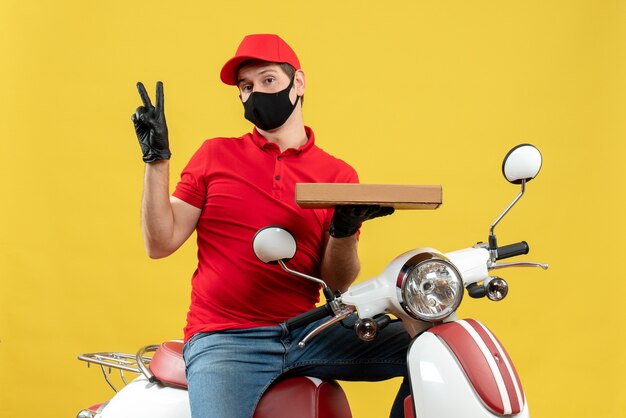 Vista superior del mensajero confundido vestido con blusa roja y guantes de sombrero en máscara médica sentado en scooter mostrando orden haciendo gesto de victoria